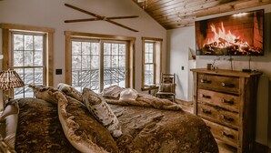 Un dormitorio acogedor con una cama grande, techo de madera y un televisor mostrando una chimenea.