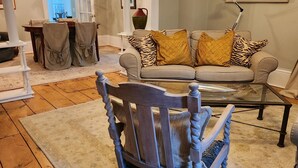 A living room with a wooden floor, a sofa with patterned cushions, a rocking chair, and a dining area with a chandelier.