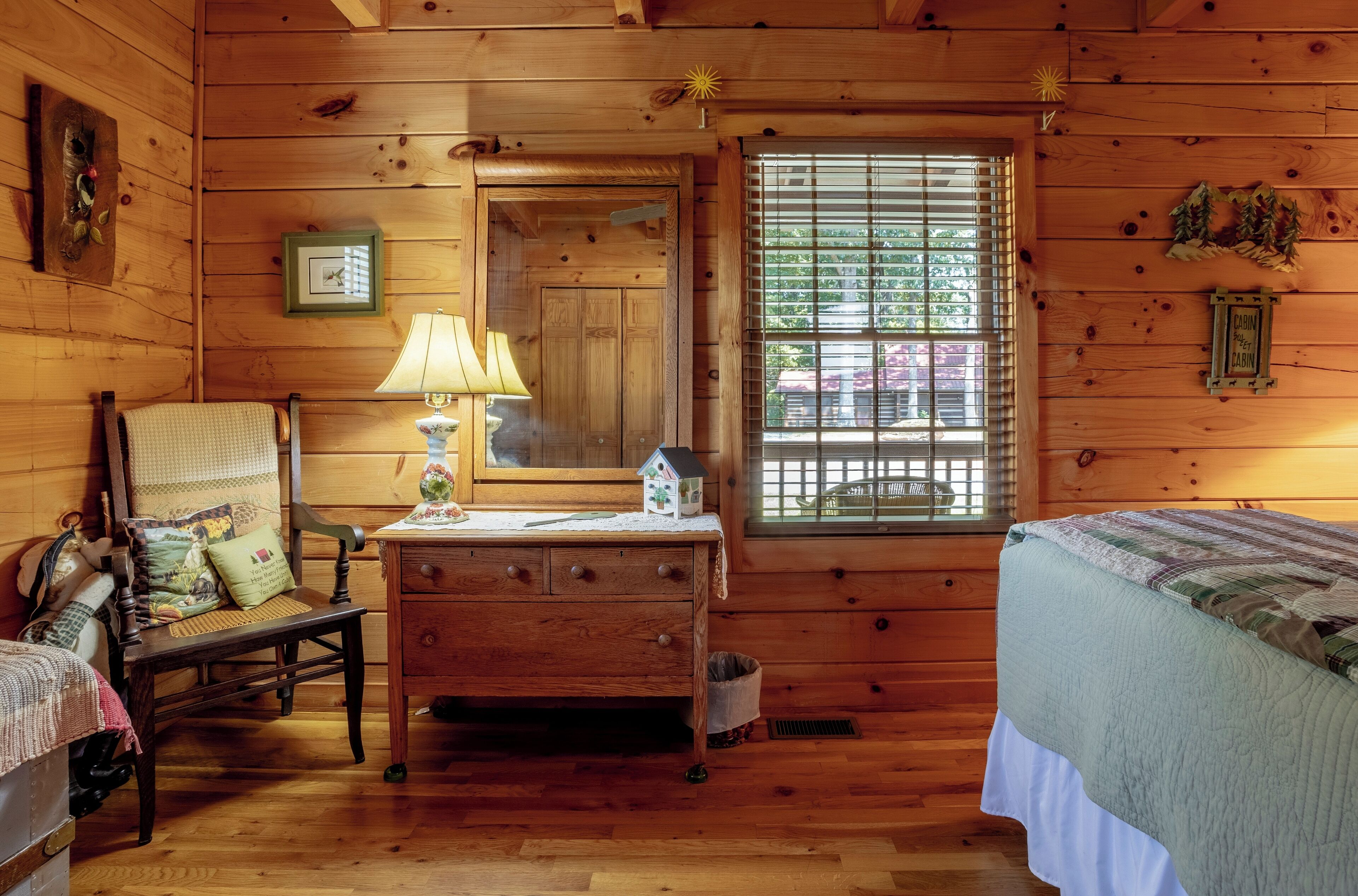 A wooden cabin room with a bed, a chair, a desk, a lamp, and a window with blinds.