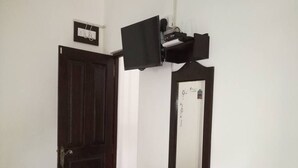 A room with a dark wooden door, a small wooden bench, a white wall, and a black shelf with a mirror and a television. - Myr-o Homestay (Anuradhapura)