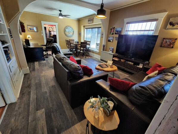 A modern living room with a sofa set, a coffee table, and a TV. There is a dining area with a table and chairs, and a kitchen area with cabinets and appliances.