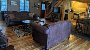 A spacious living room with a fireplace, wooden floors, and a staircase leading to a kitchen area. - Historic Lodge on 48 Acres Overlooking Long Lake Updated for Accessibility (Walker)