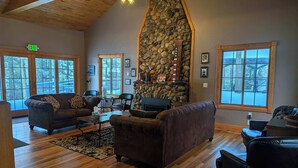 A spacious living room with a stone fireplace, wooden flooring, and large windows offering a view of the outdoors. - Historic Lodge on 48 Acres Overlooking Long Lake Updated for Accessibility (Walker)
