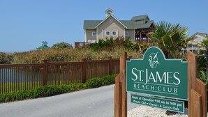 A sign for St. James Beach Club with a wooden fence and a view of a large house in the background. - Spacious Upscale Condo,  Monthly Rates Available October- April,  Call Owner (Southport)