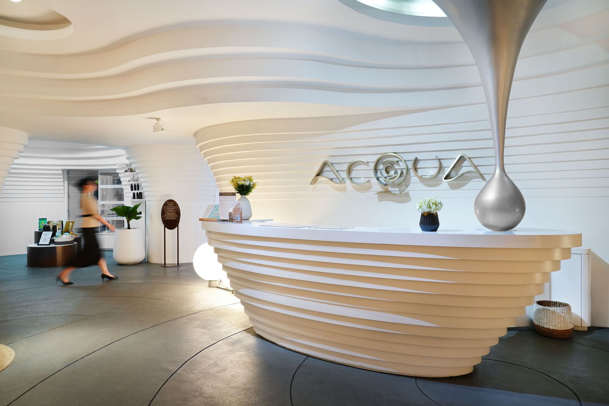 Un comptoir d’accueil moderne au design incurvé original, un grand vase métallique et une enseigne sur laquelle on peut lire « ACQUA ».