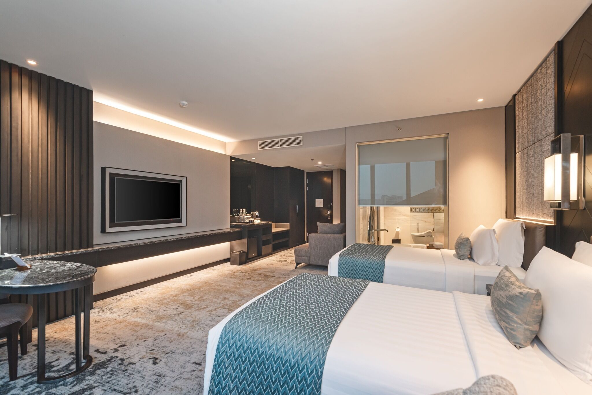A modern hotel room with a large bed, a desk, a TV, and a bathroom visible through a glass door.
