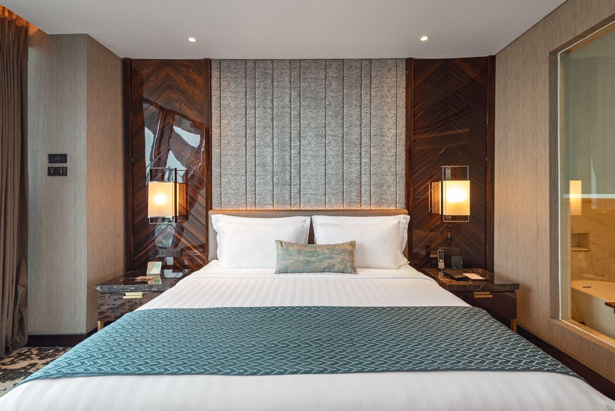 A hotel room with a large bed, two bedside tables with lamps, a wooden headboard, and a view of the bathroom.