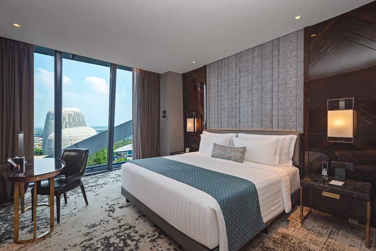 A hotel room with a large bed, a desk, a chair, and a view of a cityscape through large windows.