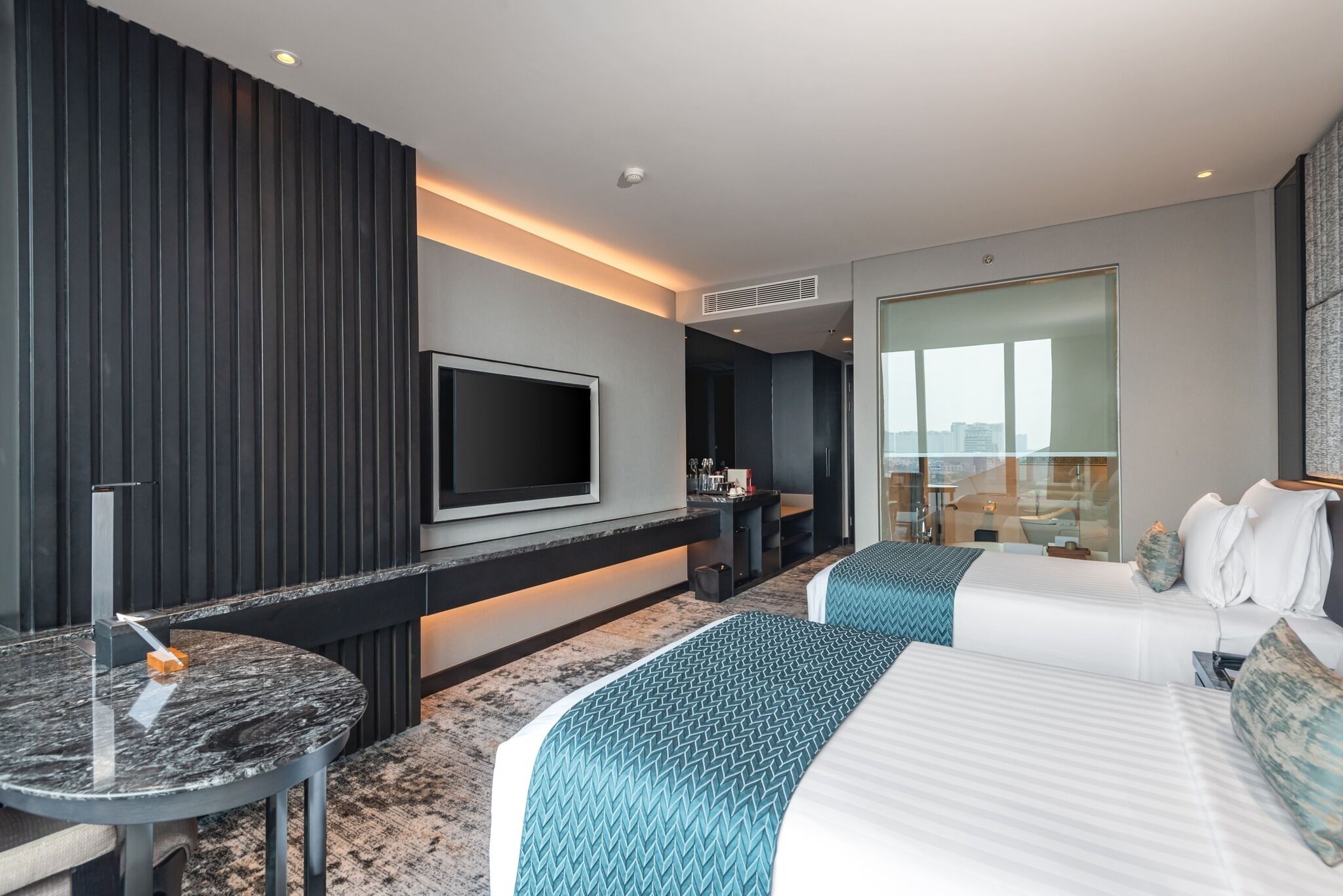 Una habitación de hotel moderna con una cama grande, un televisor montado en la pared, una mesita redonda y una zona de estar con vistas a la ciudad.