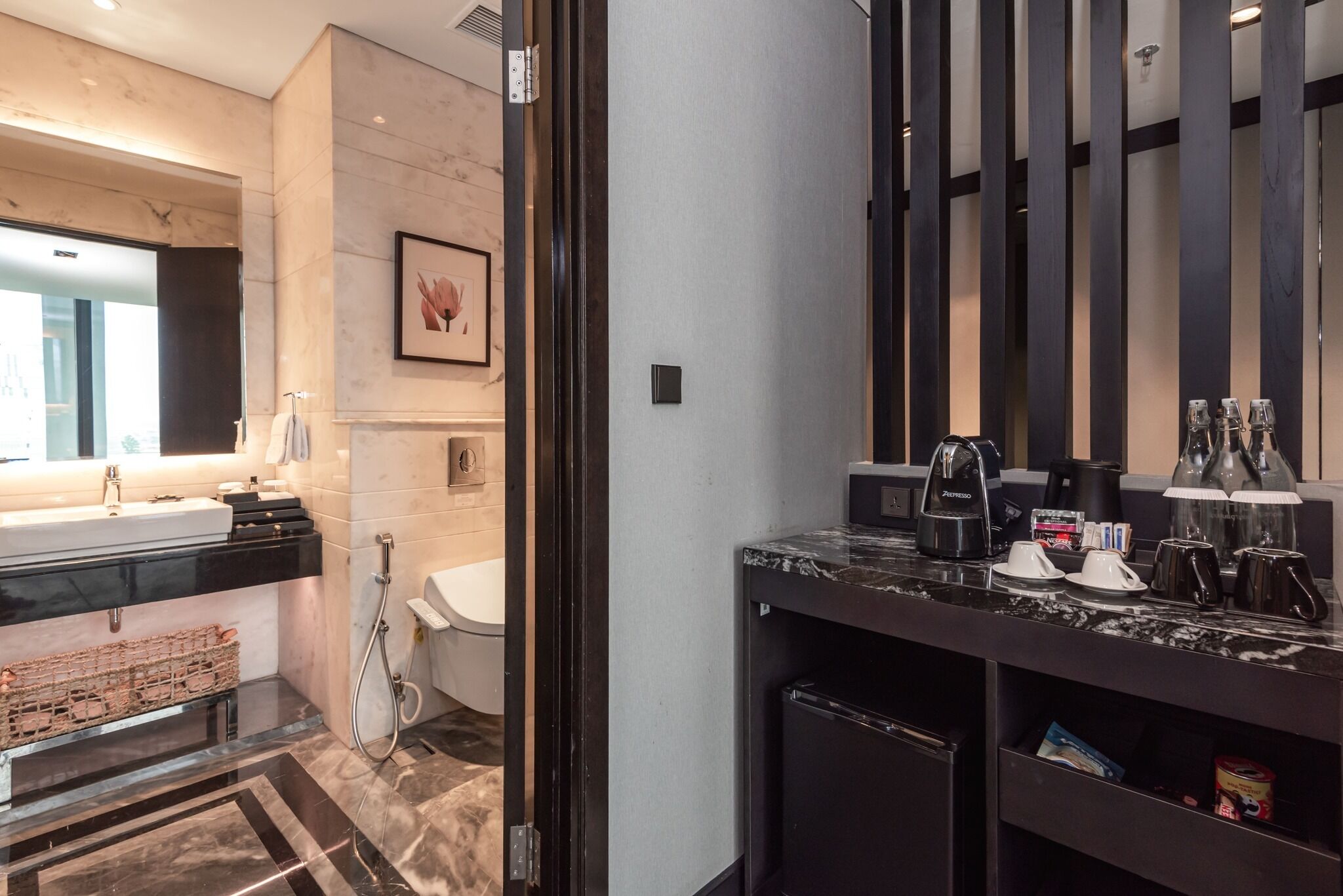 Une salle de bain moderne avec un grand lavabo, un miroir et un porte-serviettes. On y trouve également une douche avec un pommeau de douche à main et des toilettes. La salle de bain est dotée d’un sol en marbre et d’un plan de travail foncé sur lequel sont posés une machine à café et des tasses.