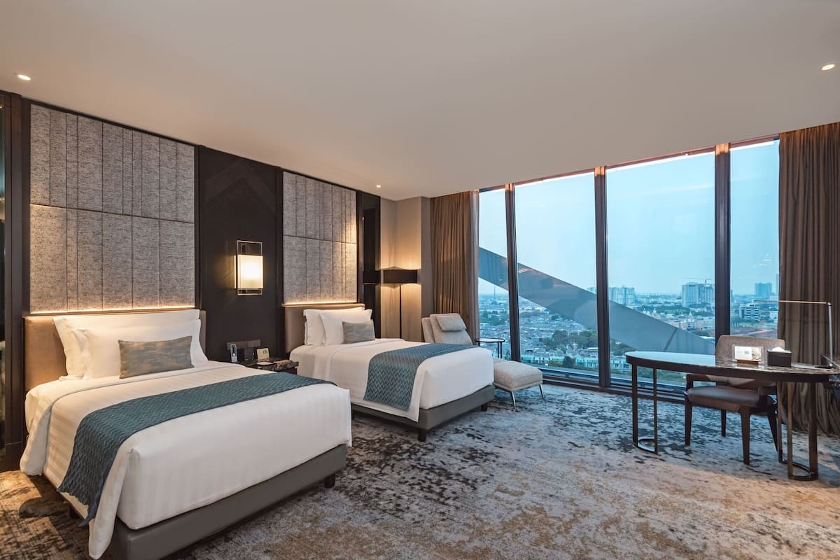 A modern hotel room with two beds, a desk, and a large window offering a city view.