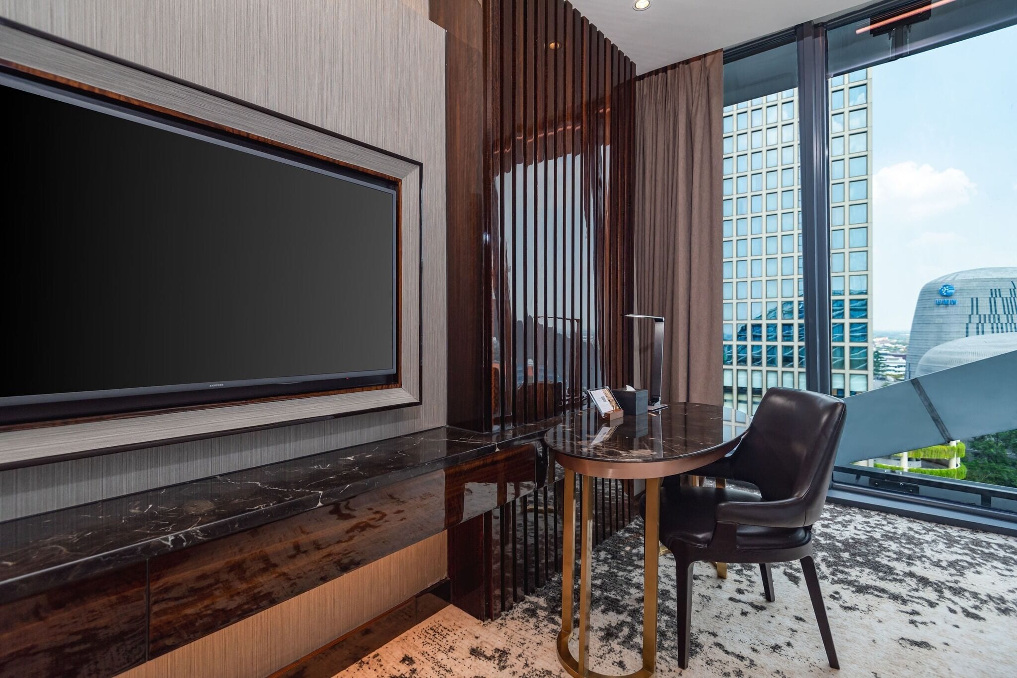 A modern hotel room with a large flat-screen TV, a round table with a chair, and a view of a cityscape through large windows.