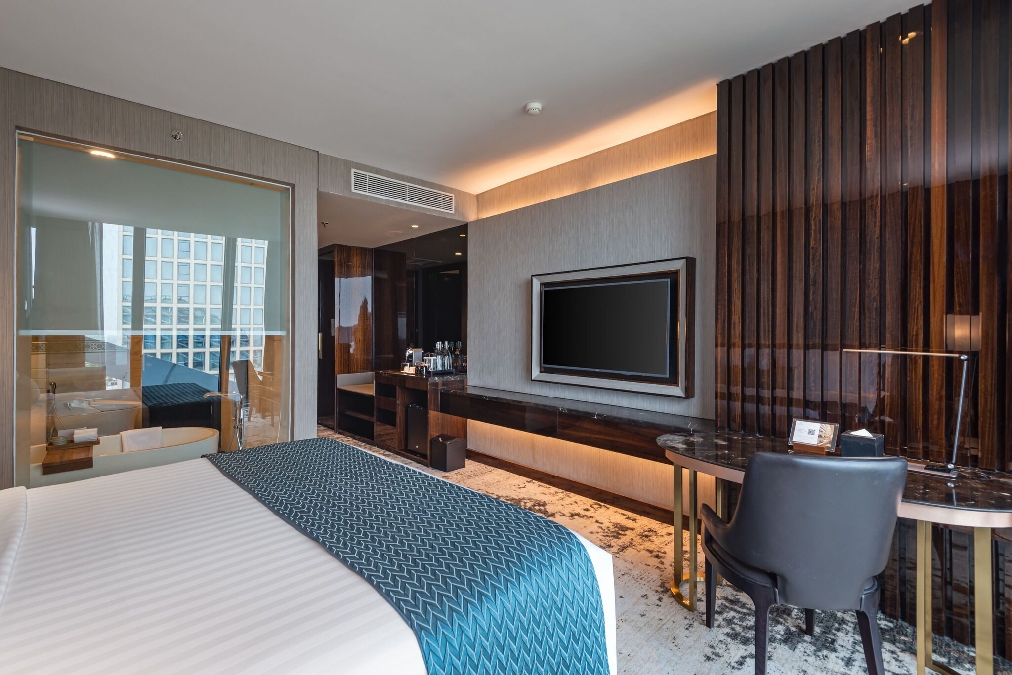 A modern hotel room with a large bed, a desk with a chair, a flat-screen TV, and a balcony with a city view.