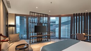 Una habitación de hotel moderna con una cama grande, zona de estar con televisor y amplias ventanas con vistas a la ciudad.