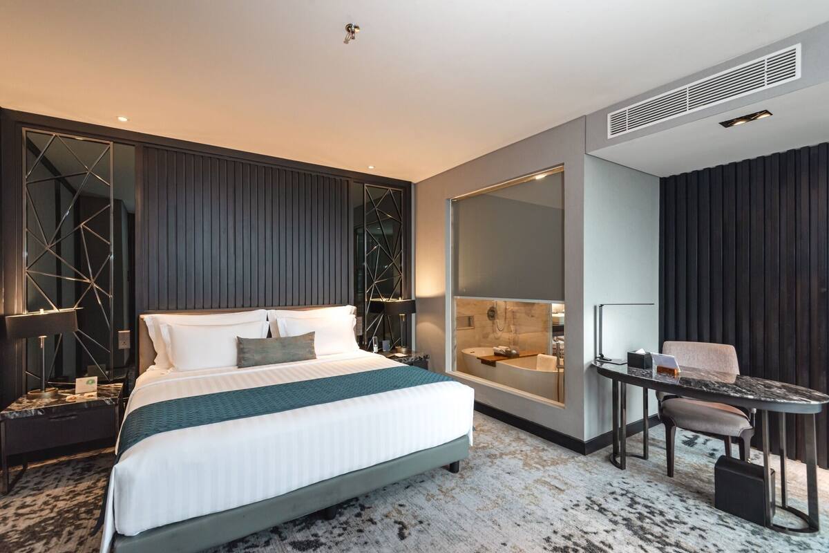 A modern hotel room with a large bed, a desk, a chair, a large mirror, and a bathroom visible through a glass partition.