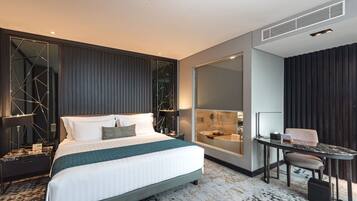 A modern hotel room with a large bed, a desk, a chair, a large mirror, and a bathroom visible through a glass partition.