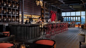 Una zona de bar con un barman, estantes con botellas y taburetes rojos.
