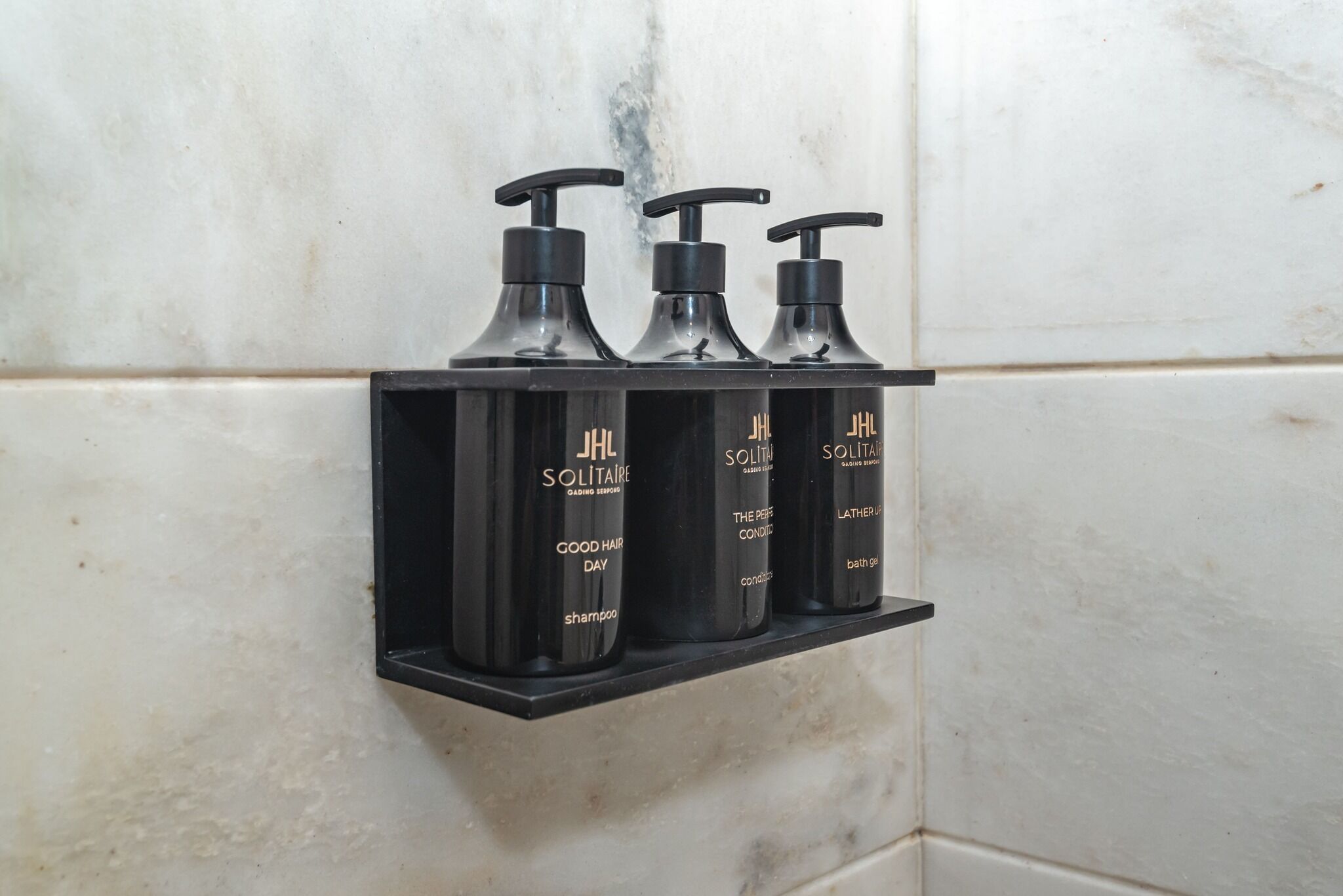Un estante negro con tres botellas negras de productos capilares JHL Solitaire, sobre un muro cubierto de azulejos blancos.