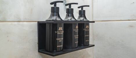Un estante negro con tres botellas negras de productos capilares JHL Solitaire, sobre un muro cubierto de azulejos blancos.