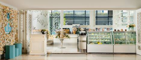 Un interior moderno con un arreglo floral central, una vitrina de pasteles y vistas al exterior con vegetación.