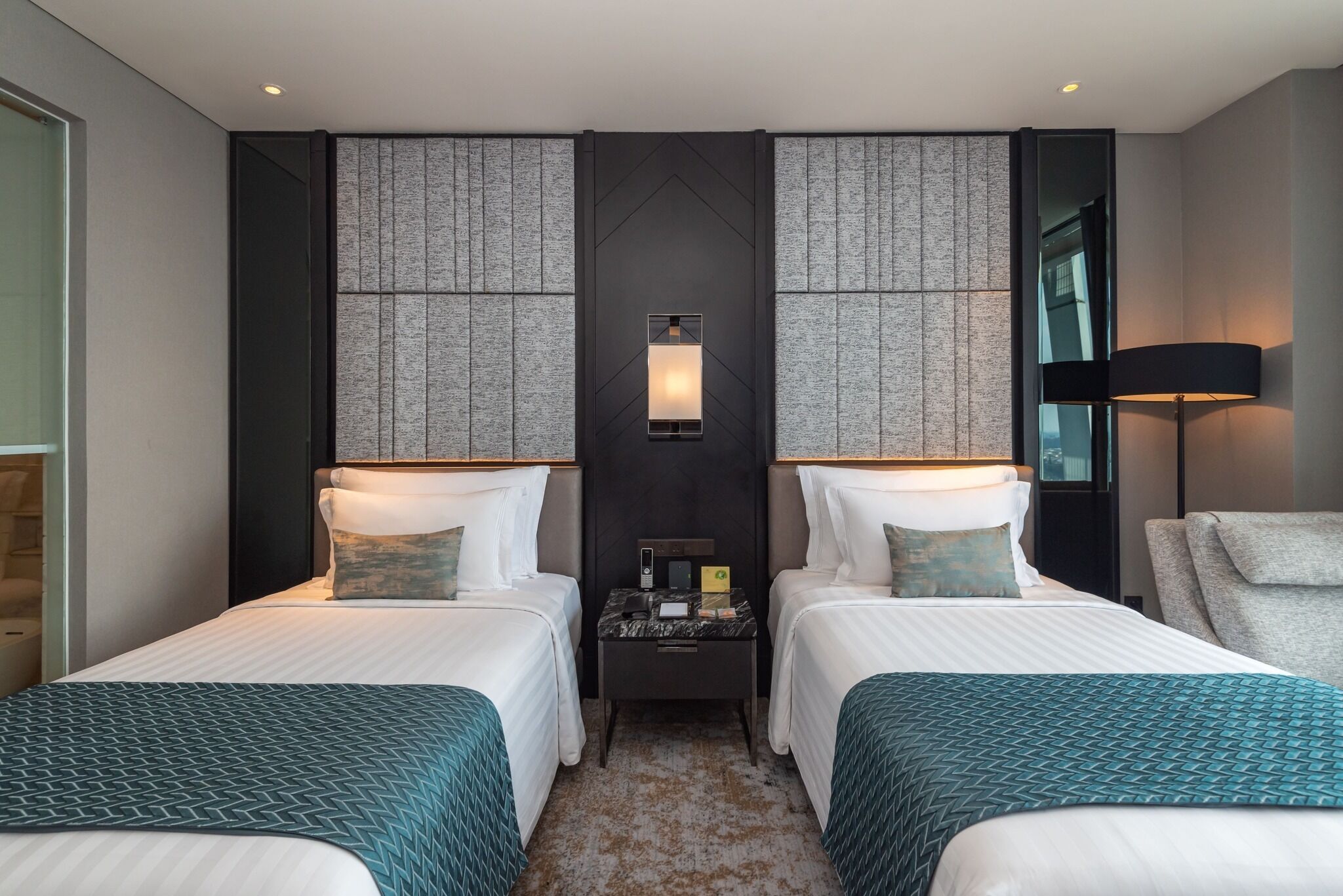 A hotel room with two beds, a nightstand, a lamp, and a view of the city.