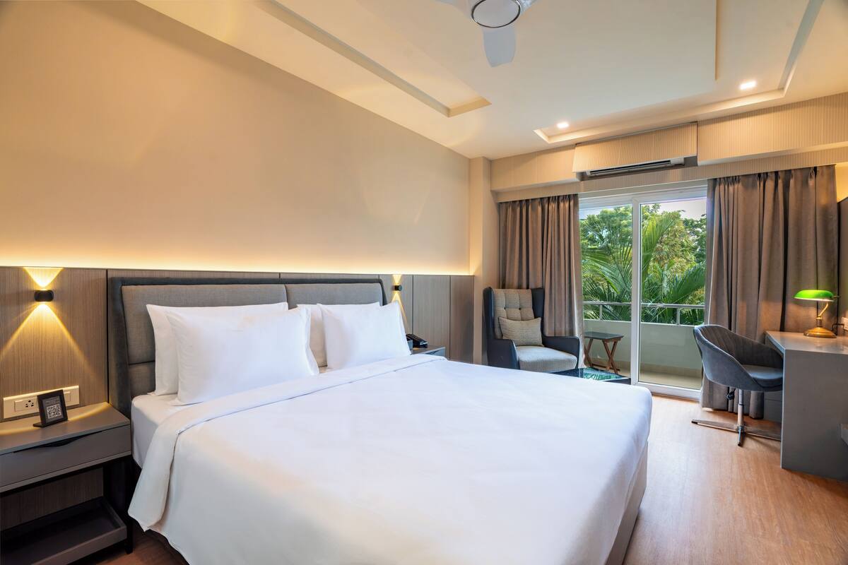Habitación de hotel con una cama grande, un escritorio con silla, una lámpara de escritorio y vistas al exterior a través de una puerta corredera.