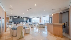Une salle de conférence spacieuse, dotée de tables et de chaises rondes, d’un comptoir d’accueil et d’une grande fenêtre donnant sur la ville.