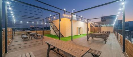 Una terraza al aire libre con mesas de picnic de madera, una escalera y un pequeño edificio con techo verde.