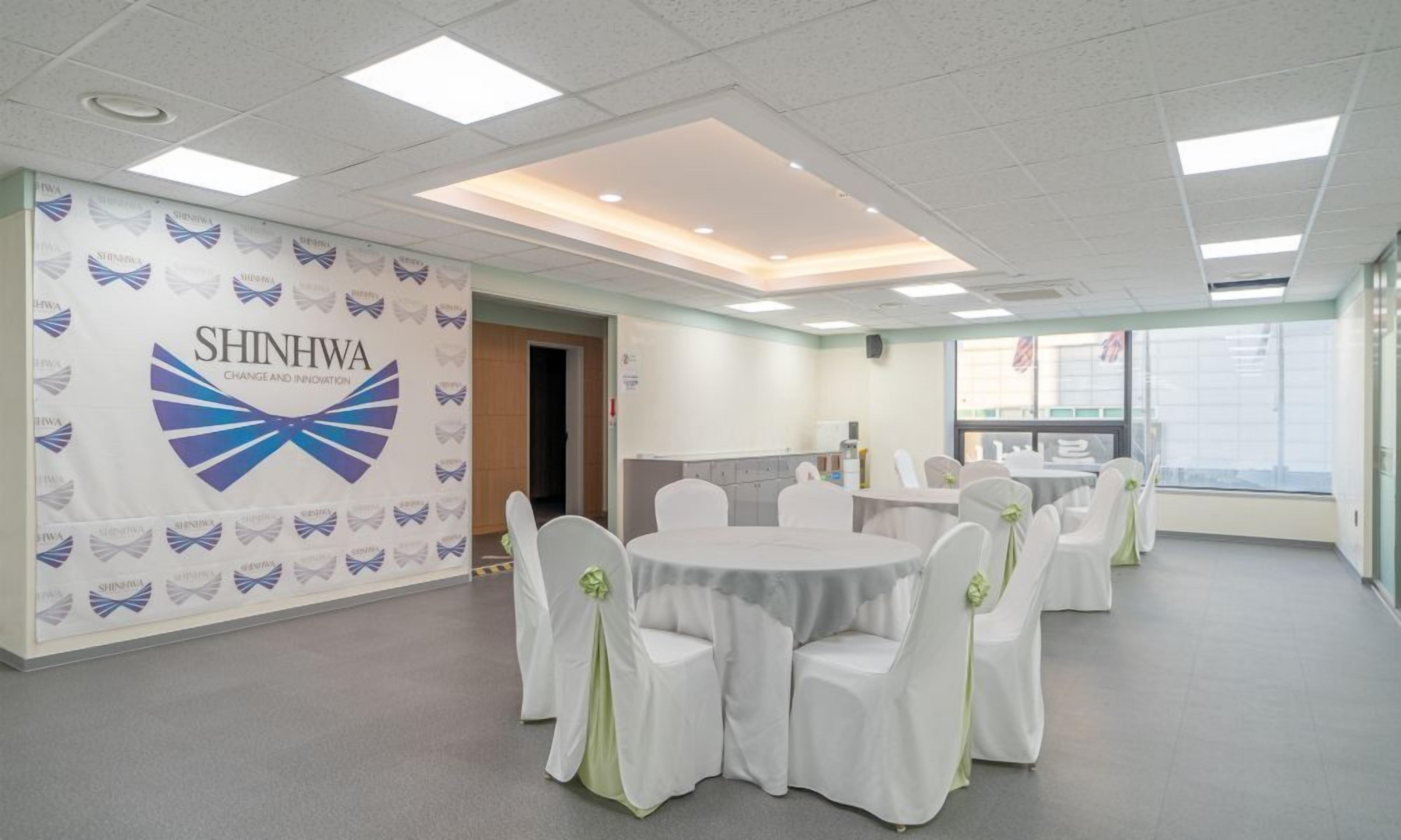 A conference room with round tables and white chairs, a banner with 'SHINNHA' branding, and a view of the outside through a window.