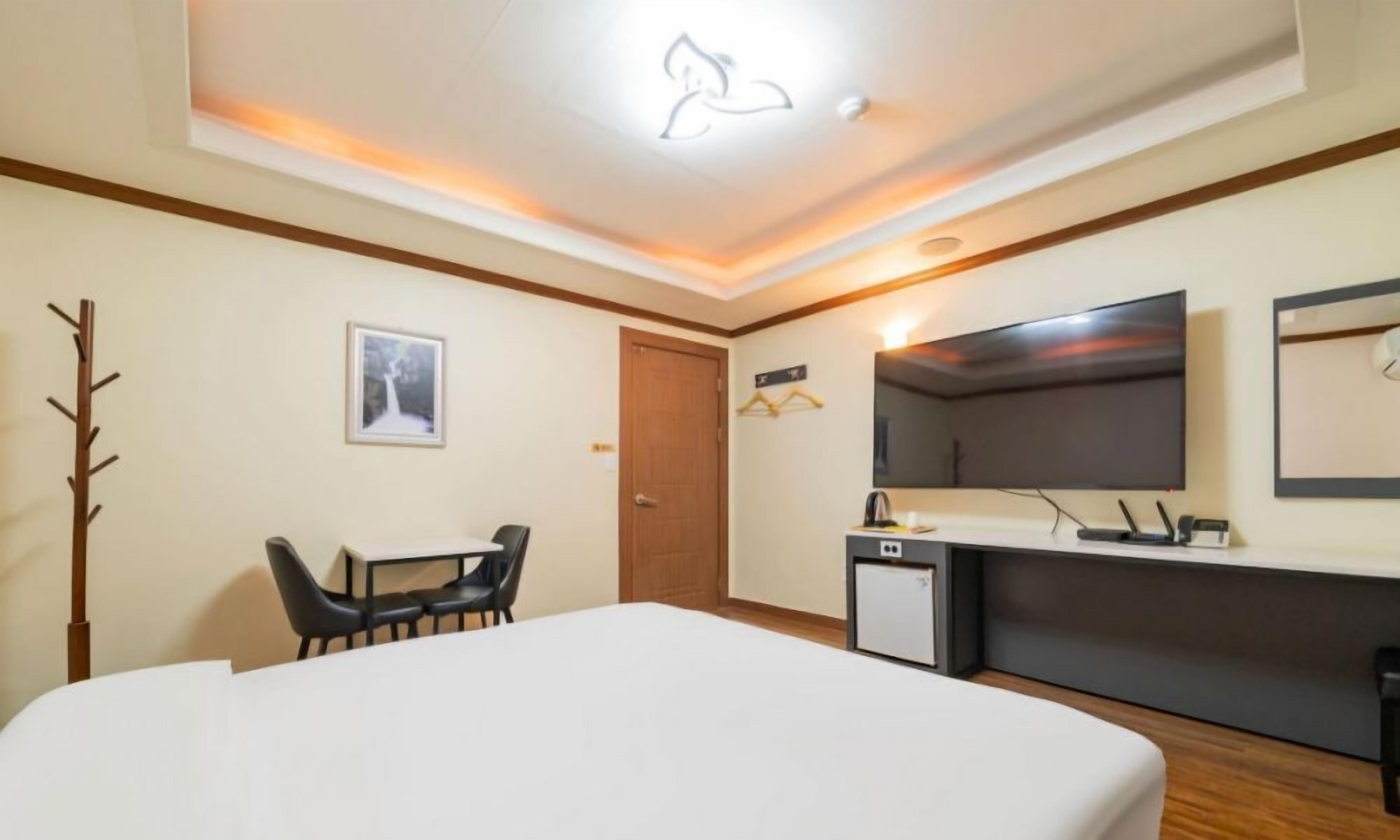 A hotel room with a bed, a desk, a chair, a small kitchenette, a TV, and a coat rack.