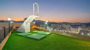 Une terrasse sur le toit, avec un revêtement d’herbe artificielle, un escalier blanc et une vue sur la ville au crépuscule.