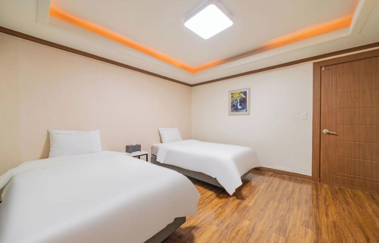 Een hotelkamer met twee bedden, een houten vloer, een plafondlamp en een deur.