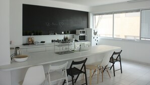 A modern kitchen with a white countertop, a blackboard with chalk writing, and a window with blinds. - TLV Suites on the beach (Tel Aviv)