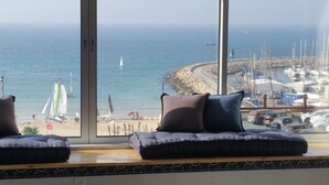 A view from a window overlooking a beach with sailboats, a marina, and a rocky shoreline. - TLV Suites on the beach (Tel Aviv)