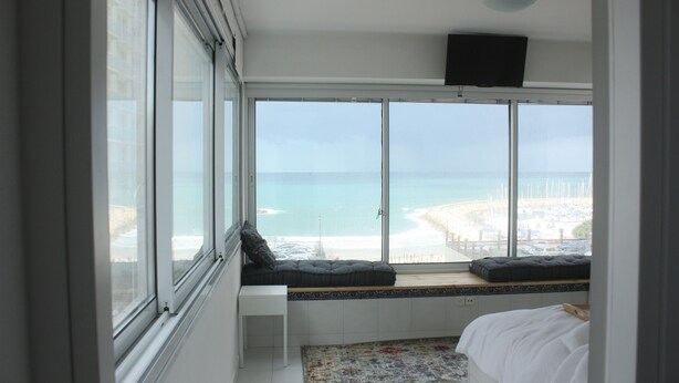 A bedroom with a large window overlooking a beach, a bed, a small white nightstand, and a television mounted on the wall. - TLV Suites on the beach (Tel Aviv)