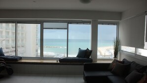 A modern living room with a large window overlooking a beach and high-rise buildings. - TLV Suites on the beach (Tel Aviv)