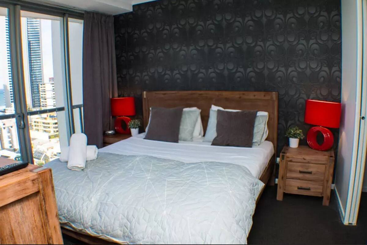 A modern hotel room with a large bed, two bedside tables with lamps, a view of the city through large windows, and a patterned wallpaper on the wall.
