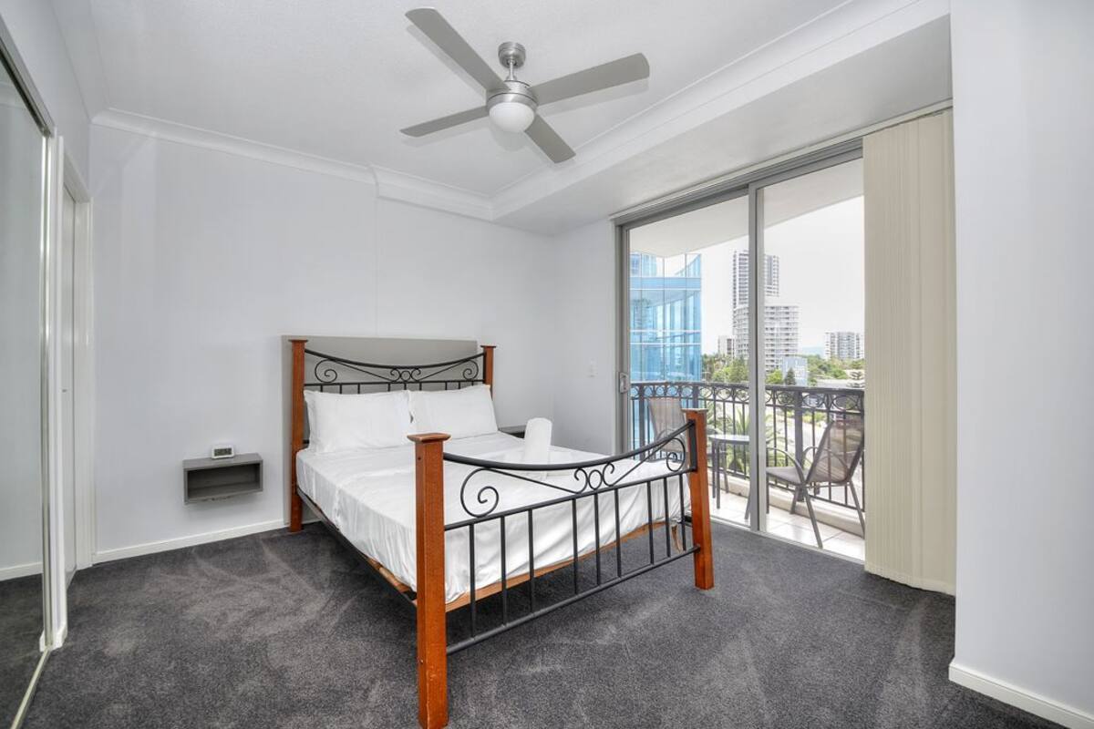 A bedroom with a bed, a ceiling fan, a window with a view of buildings, and a balcony with chairs.