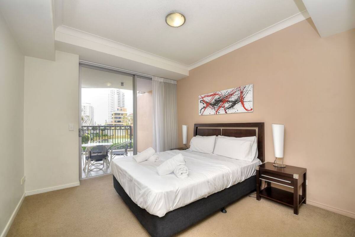 A hotel room with a bed, a nightstand, a painting, and a balcony with a view of buildings.