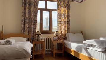 Een hotelkamer met twee houten bedden, een raam met gordijnen, een radiator en een lamp op een nachtkastje van hout.