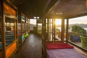 A dormitory room with bunk beds, a balcony with a view of the mountains, and a large window allowing sunlight to enter. - One Tree Hill - Hostel (Labuan Bajo)