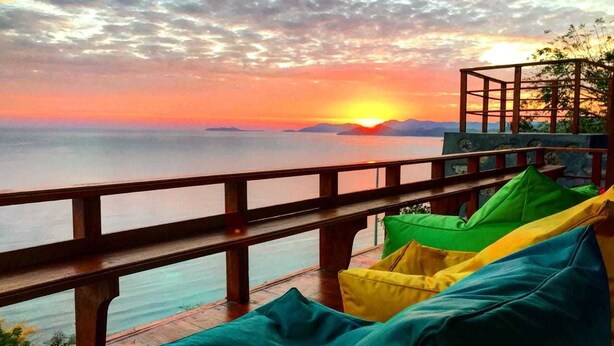 A sunset view from a balcony with colorful cushions and a wooden railing overlooking the sea. - One Tree Hill - Hostel (Labuan Bajo)