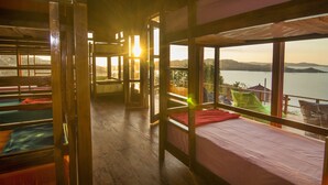 A cabin with bunk beds, wooden floors, and large windows offering a view of a lake and mountains. - One Tree Hill - Hostel (Labuan Bajo)