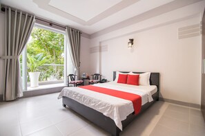 Quarto de hotel com uma cama grande, uma janela com cortinas, uma mesa pequena com uma cadeira e um vaso de plantas.