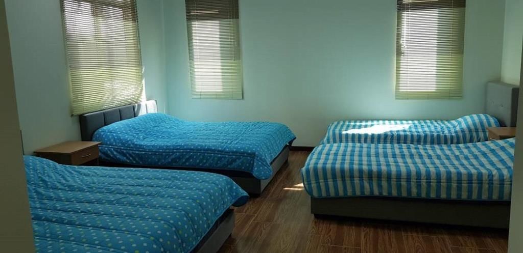 Quarto de estudante com três camas, cada uma com um edredom de padrão diferente. Há duas janelas com persianas e o chão é de madeira.