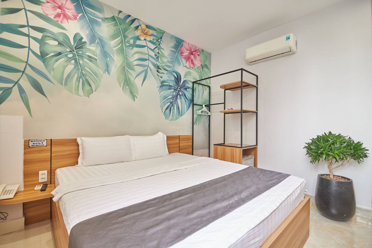 Une chambre d’hôtel moderne avec un grand lit, une fresque aux motifs tropicaux, une plante en pot et une bibliothèque intégrée.