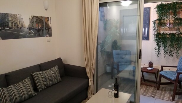A living room with a dark sofa, a glass door leading to a balcony, and a table with a bottle and glasses. - TLV Suites By The Sea - 2 Rooms (Tel Aviv)