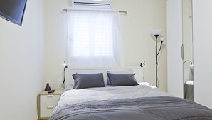 A bedroom with a bed, bedside table, lamp, and air conditioning unit. - TLV Suites By The Sea - 2 Rooms (Tel Aviv)