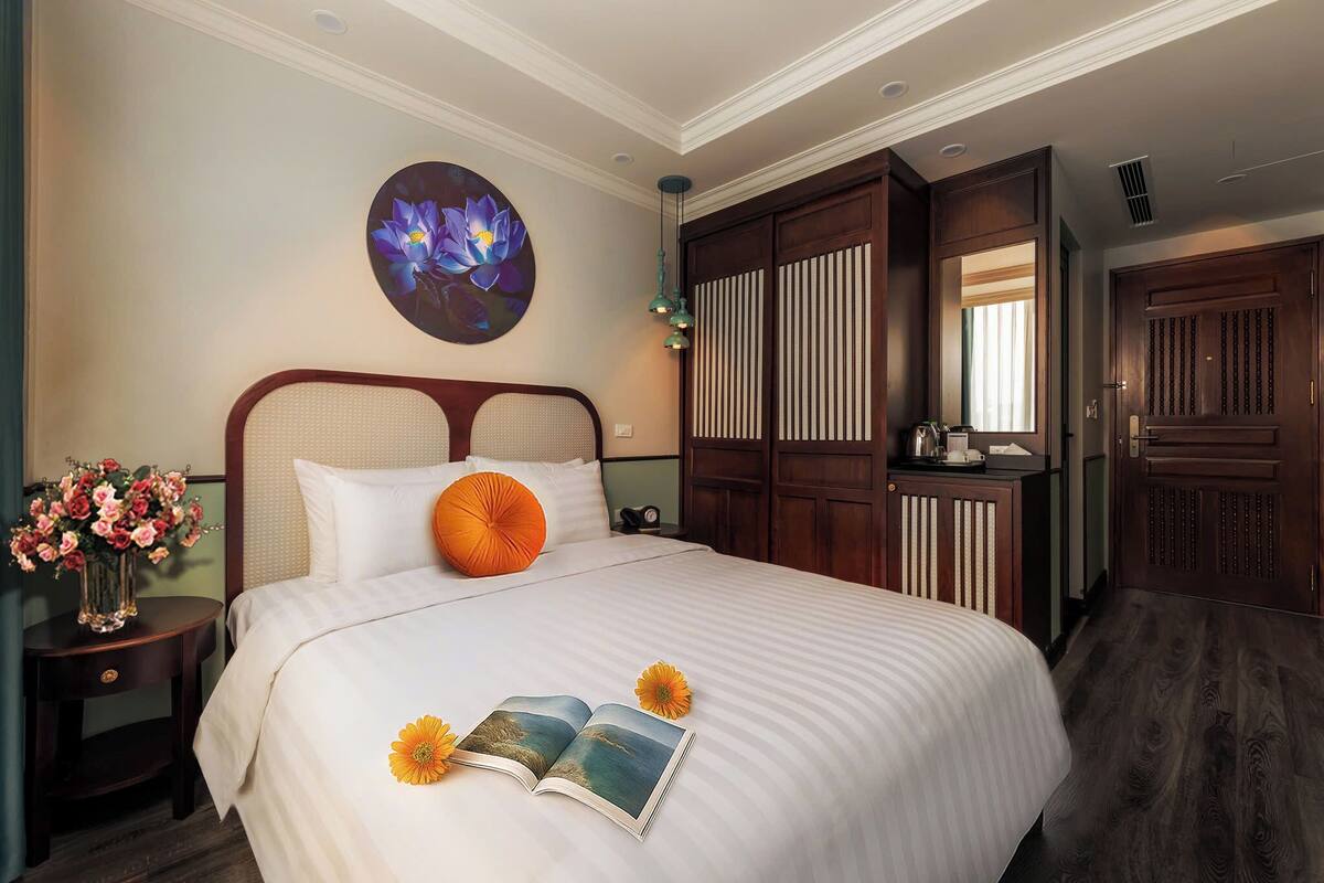 A neatly made bed with a book and flowers, a vase of flowers, and a painting on the wall.