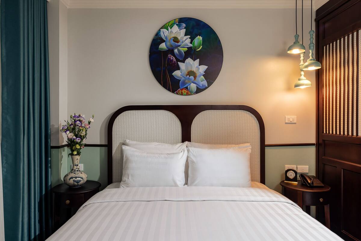 A neatly made bed with white linens, a round wall decoration of flowers, a vase with flowers, and a small table with a clock and a phone.